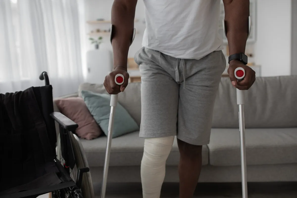 A person with a bandaged leg using crutches near a wheelchair in a living room setting.