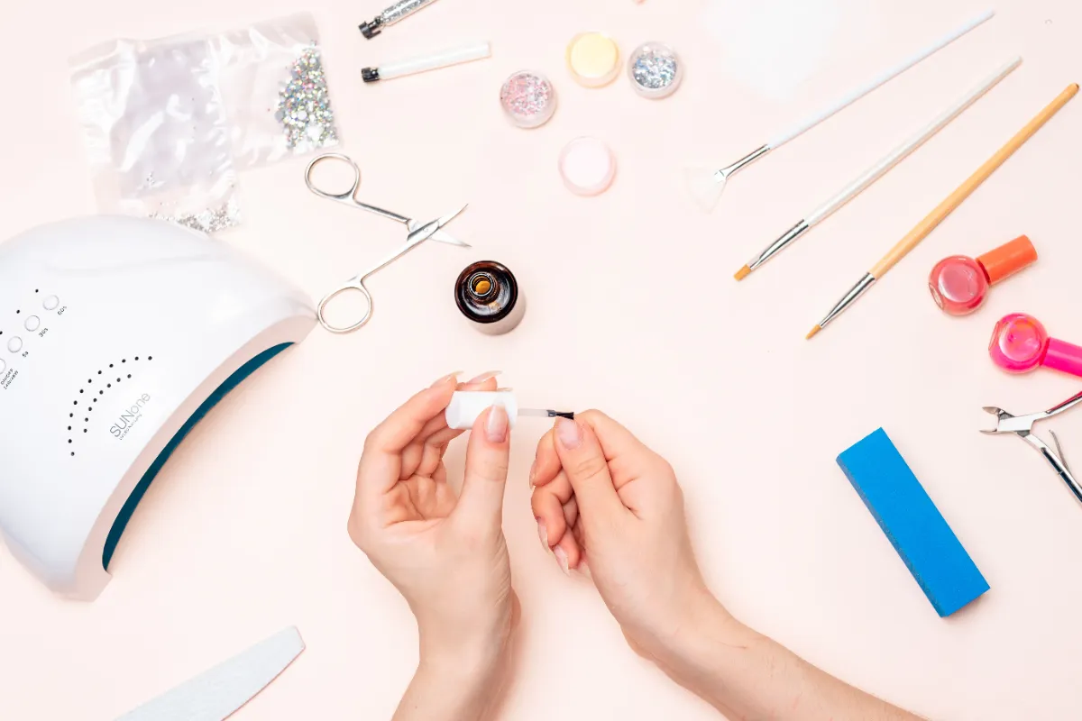 Hands painting nails with clear polish surrounded by nail art supplies, including a UV lamp, brushes, glitter, and nail files on a light pink surface.