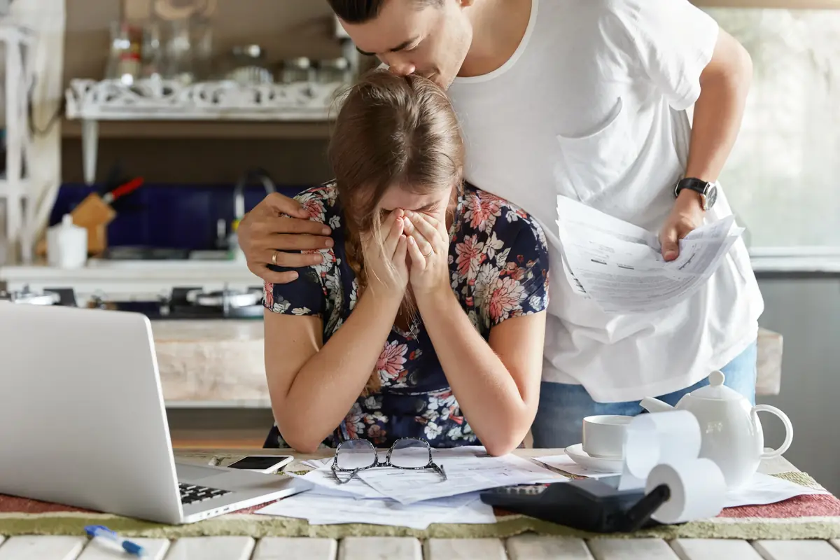 A woman sits at a table overwhelmed by bills and paperwork, with a laptop and calculator nearby, while a man offers comfort by embracing and kissing her head.