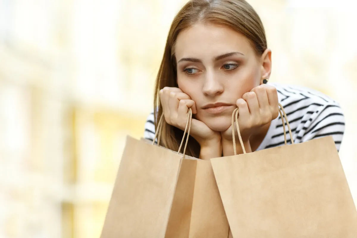 A woman with a contemplative expression rests her chin on her hands while holding shopping bags, suggesting introspection after a purchase.