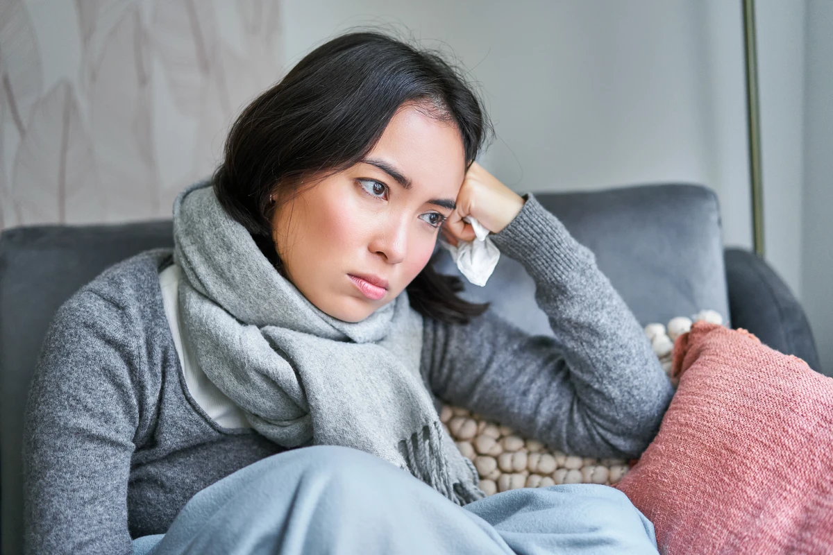Woman sitting on a couch looking pensive and tired, wrapped in a scarf with a tissue in hand, leaning against a cushion.