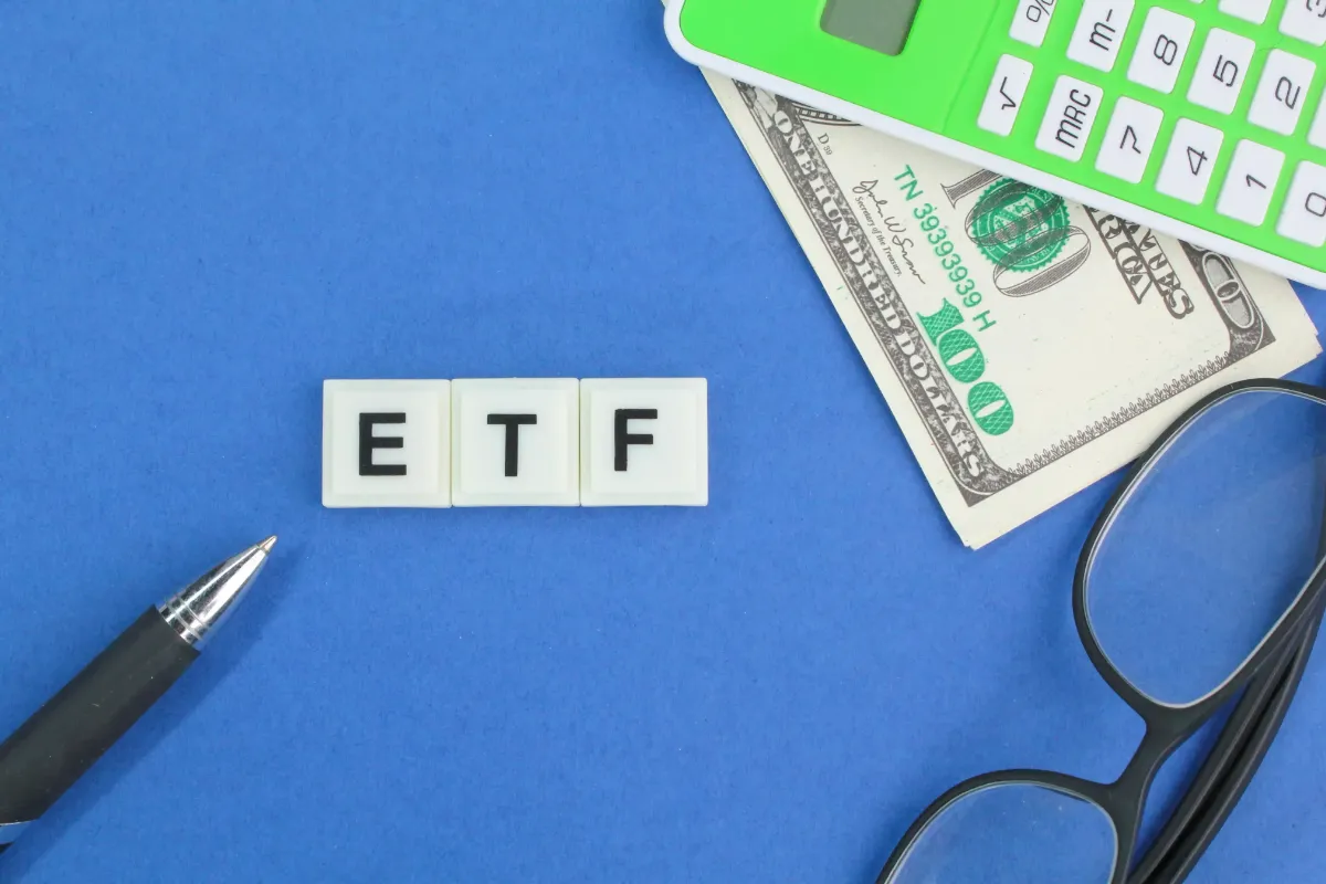 Scrabble tiles spelling "ETF" on a blue background surrounded by a pen, a pair of eyeglasses, a green calculator, and a hundred-dollar bill.