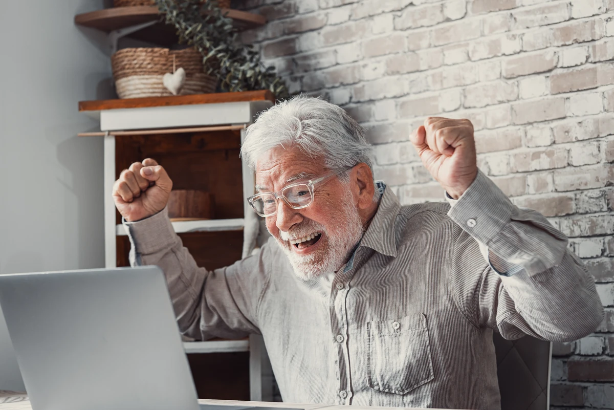 Elderly man with glasses smiling and raising his fists in excitement while looking at a laptop.
