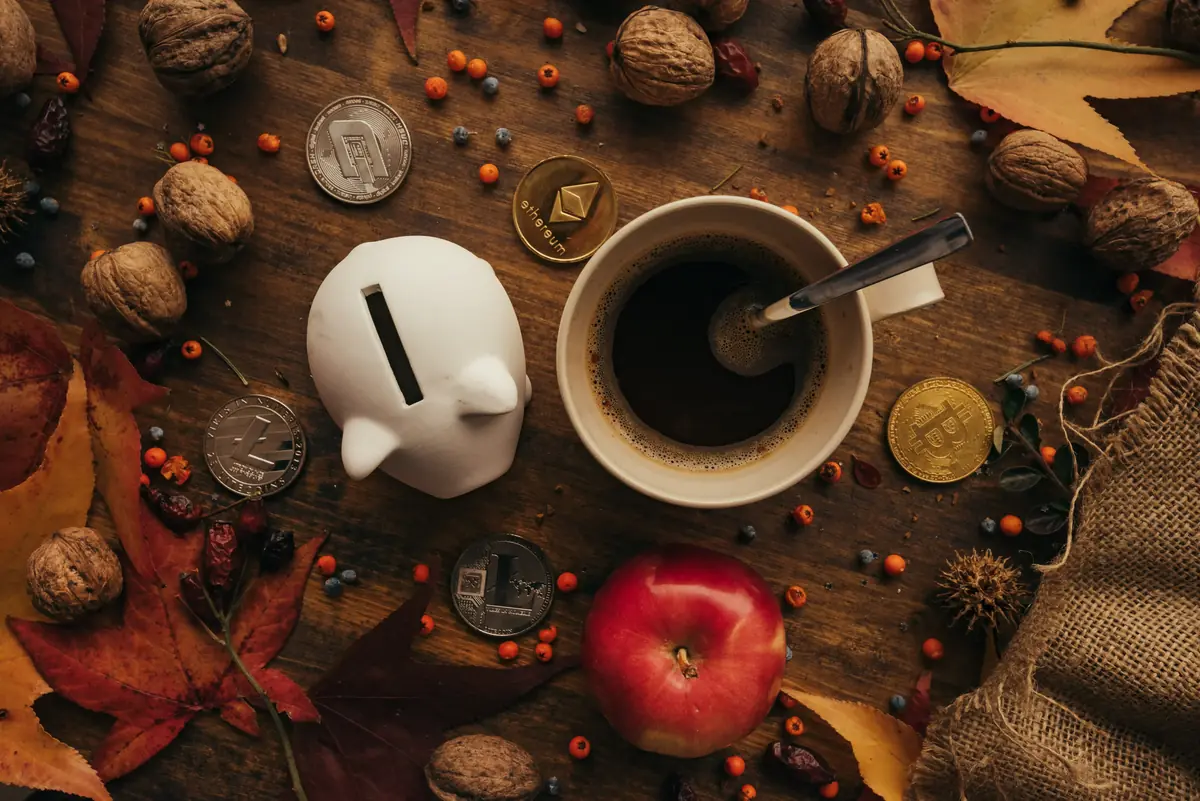 Coins and a white piggy bank alongside a coffee cup surrounded by autumn leaves, walnuts, and an apple on a wooden table.