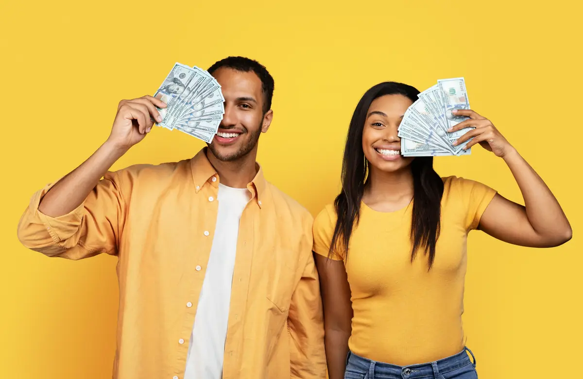 A smiling couple in casual yellow clothing holds up fans of USD notes against a matching yellow background.