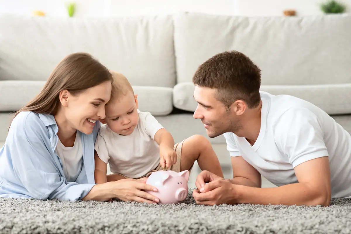 A family of three, including a mother, father, and toddler, lying on a carpet and interacting with a pink piggy bank, suggesting a focus on saving or financial planning.