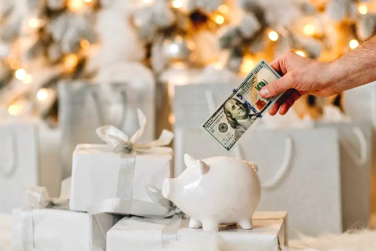 Hand placing a $100 bill into a white piggy bank in front of festive, wrapped gifts with a sparkling Christmas tree in the background.