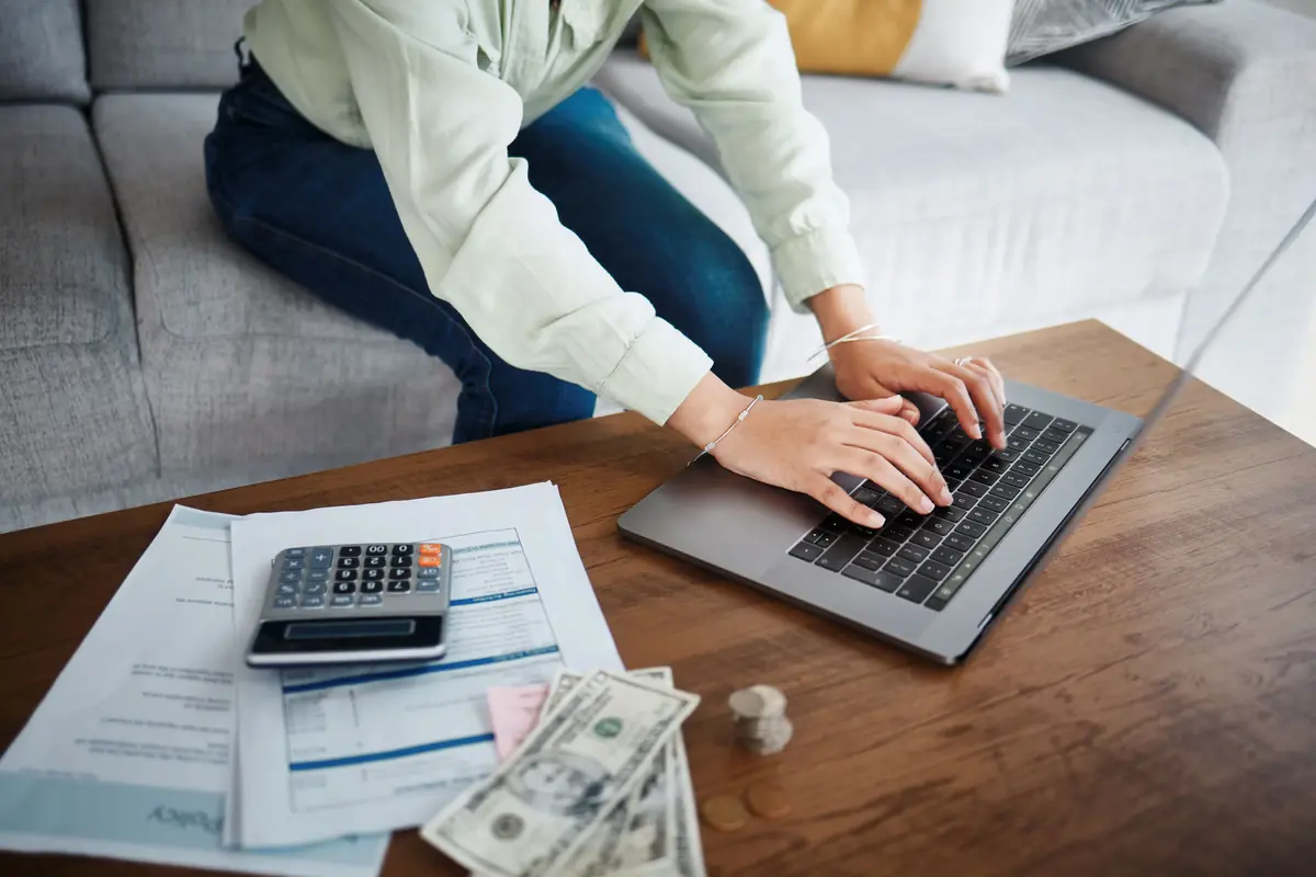Person typing on a laptop at a wooden table with scattered bills, a calculator, and cash, indicating financial planning or budgeting.
