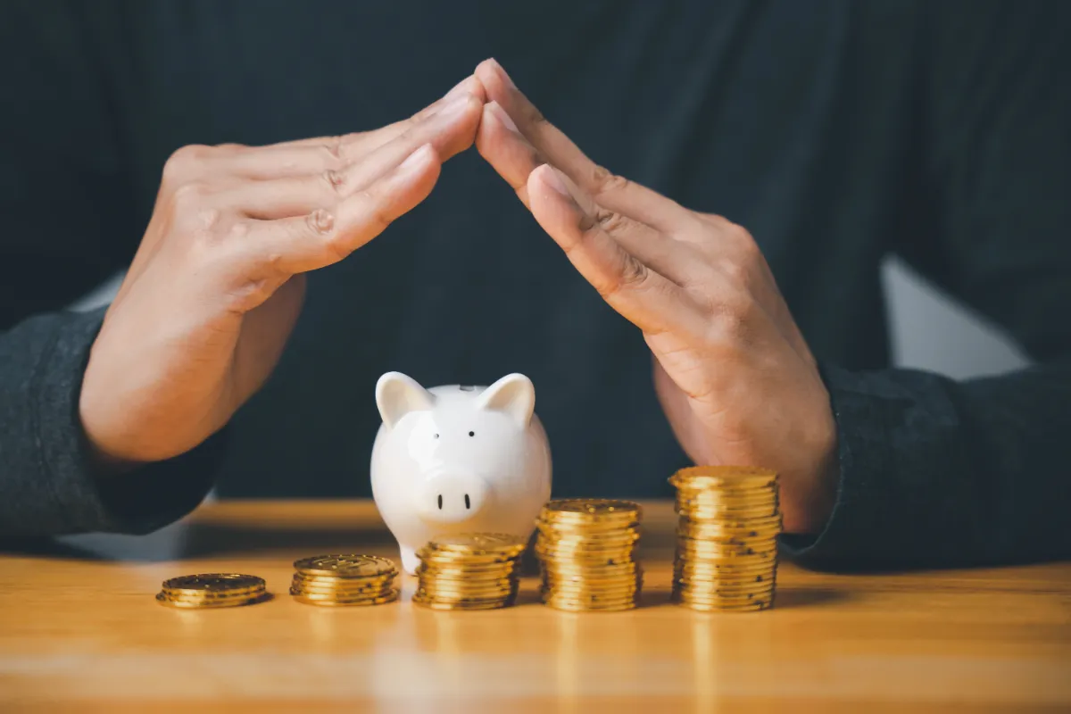Hands forming a protective gesture over a white piggy bank surrounded by stacks of gold coins on a wooden surface.