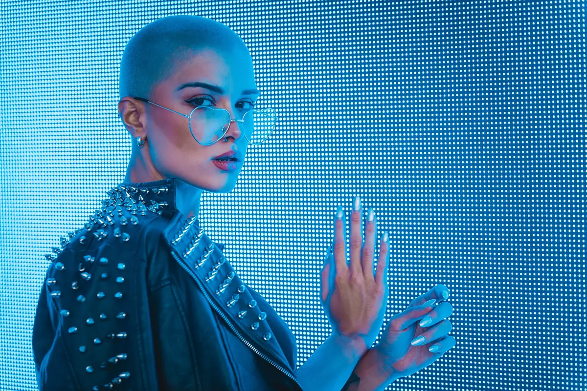 Person with buzzed hair wearing aviator sunglasses and a studded leather jacket, posing against a blue LED backdrop with long, futuristic nails.