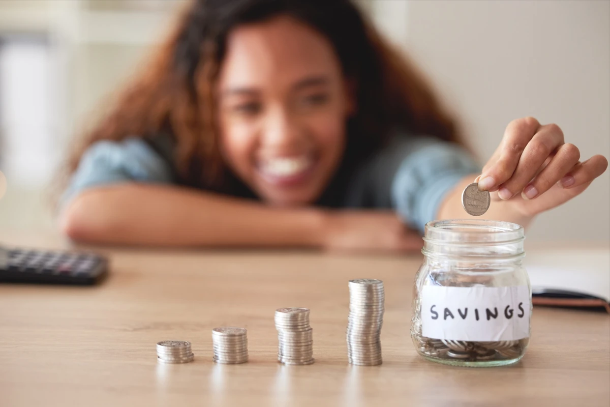 Woman adding coin to savings jar, stacks of coins nearby