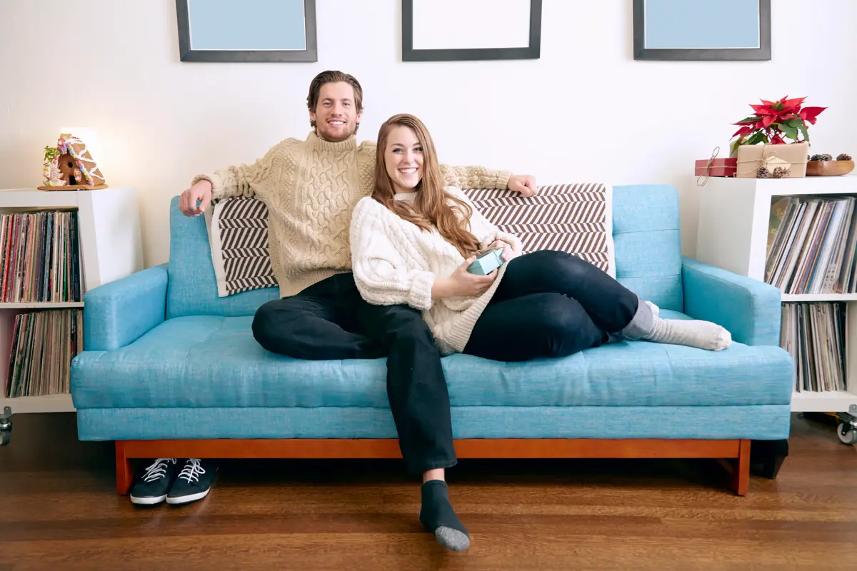 A smiling couple sitting on a blue couch in a cozy living room with framed pictures, a record collection, and a poinsettia plant in the background.