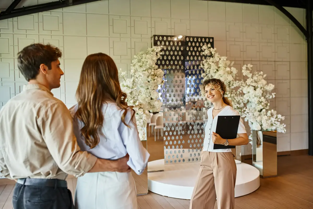 Event manager with a clipboard smiles while showing a couple a wedding venue adorned with white floral arrangements and reflective decor.