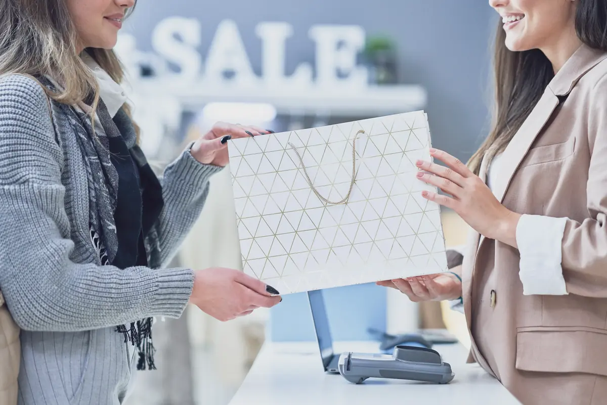 Two women are smiling while exchanging a shopping bag across a checkout counter, with the word “SALE” visible in the background.