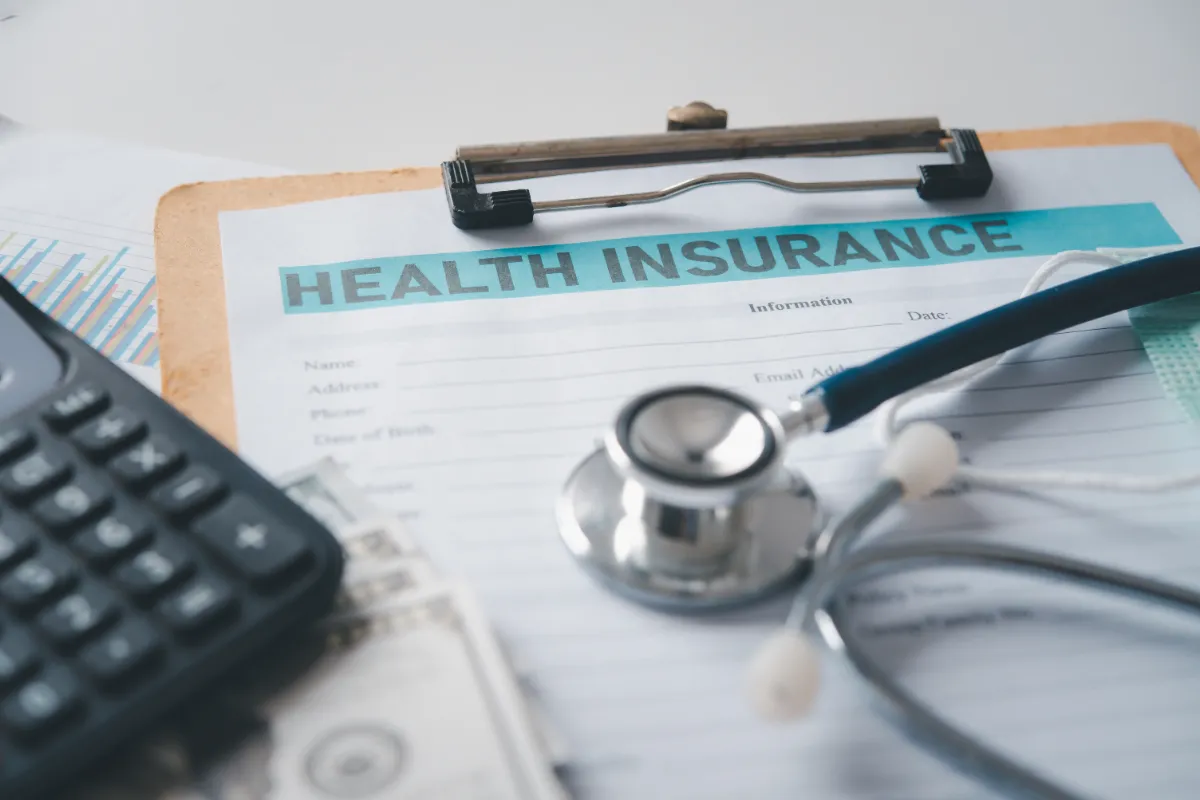 A health insurance form on a clipboard, accompanied by a stethoscope, a calculator, and a stack of dollar bills.