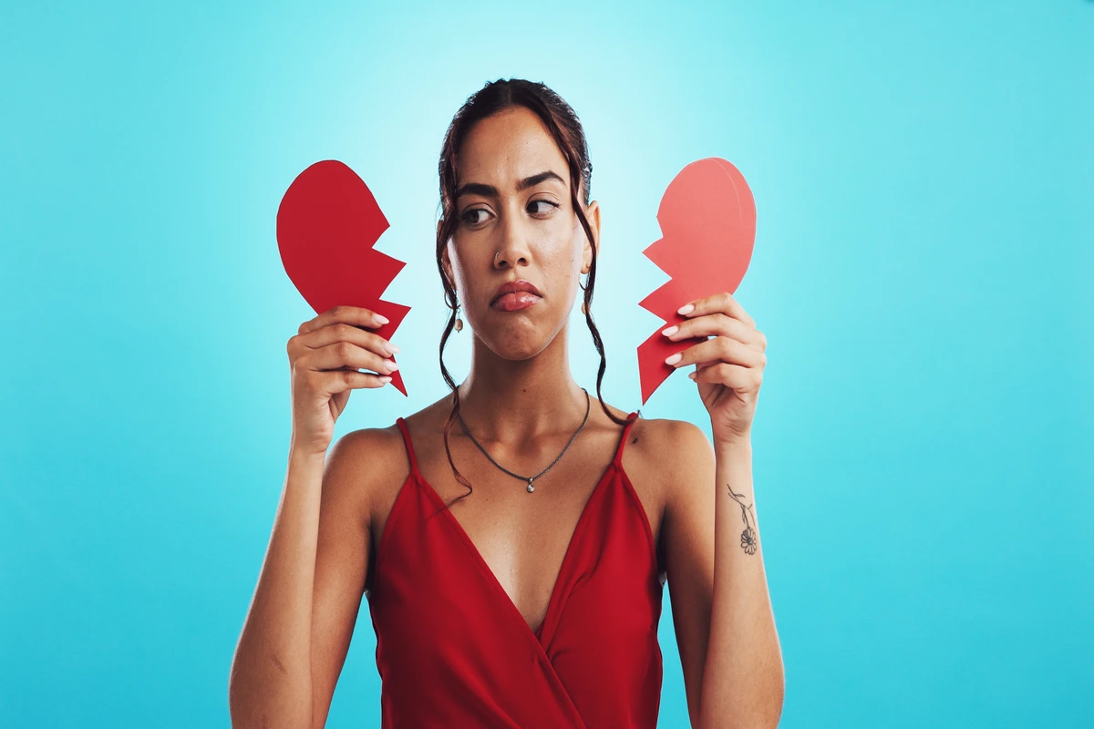A woman in a red dress holds two halves of a broken red heart with a sad expression against a blue background.