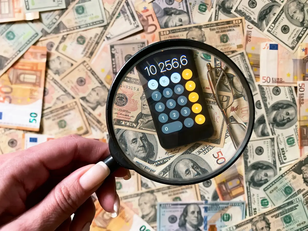 A magnifying glass focuses on a smartphone with a calculator app displaying numbers, surrounded by various denominations of U.S. dollar bills.