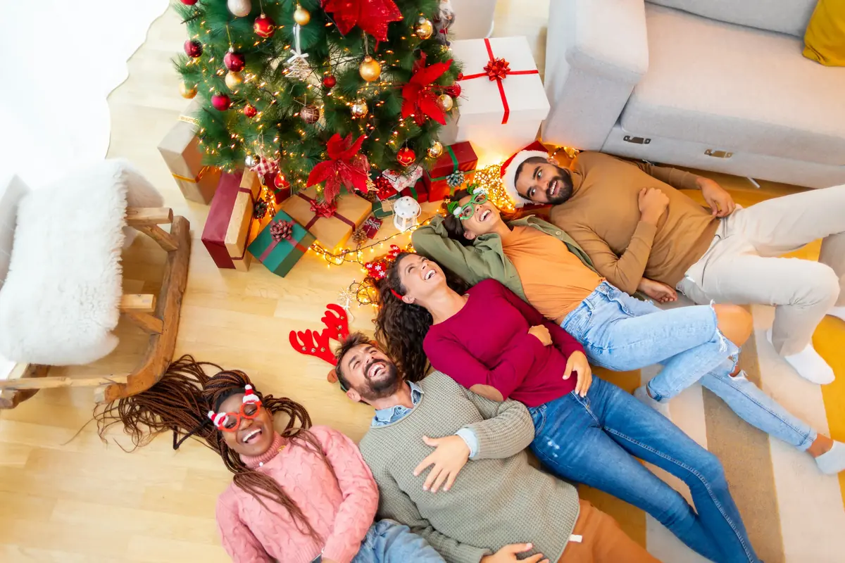 Five friends lie joyfully on the floor under a decorated Christmas tree surrounded by wrapped gifts, wearing festive accessories like reindeer antlers and red glasses.