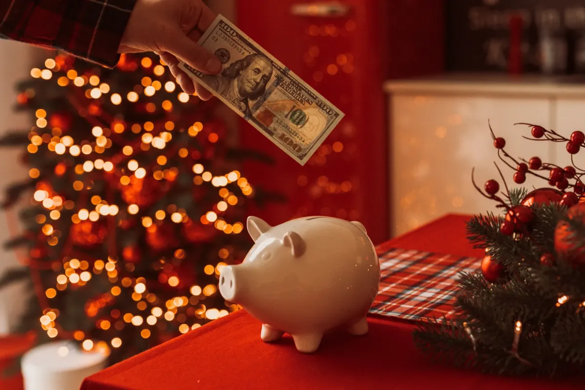 A hand holding a hundred-dollar bill above a white piggy bank on a red table, with a decorated Christmas tree and festive lights in the background.