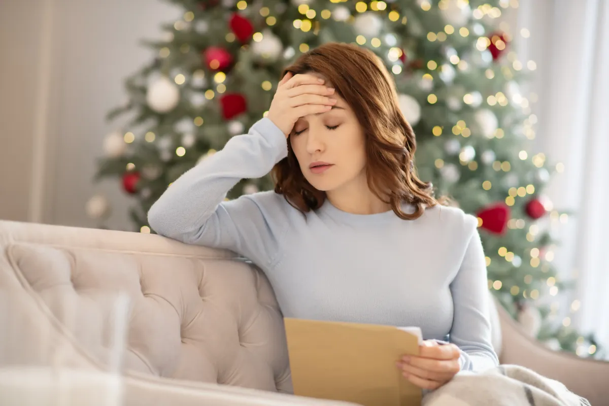 A woman sitting on a couch with her hand on her forehead, looking stressed, in front of a decorated Christmas tree while holding an envelope.
