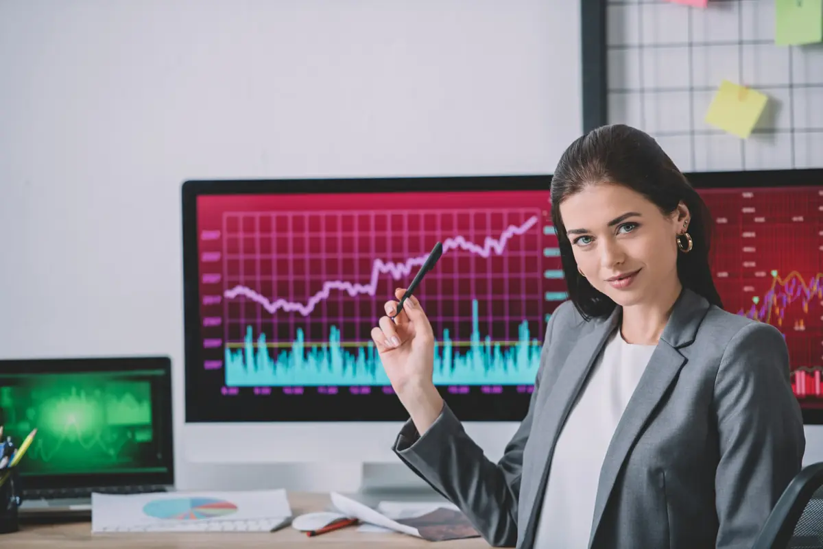 A confident woman in a gray blazer holds a pen while sitting at a desk with multiple computer screens displaying stock market charts.