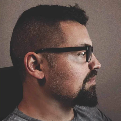 A person with a beard, glasses, and a short haircut, sitting in profile against a neutral background.
