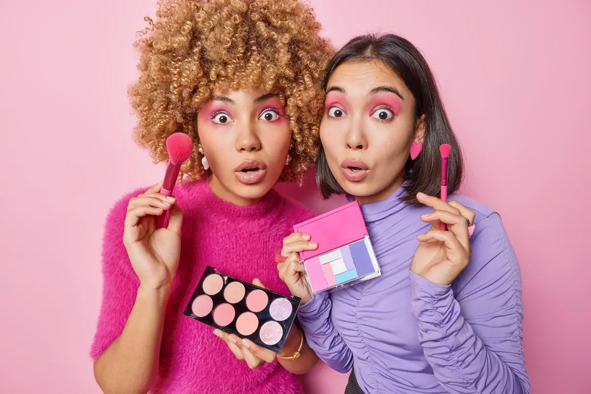 Two surprised women wearing bright, colorful makeup and holding various blush and eyeshadow palettes against a pink background.