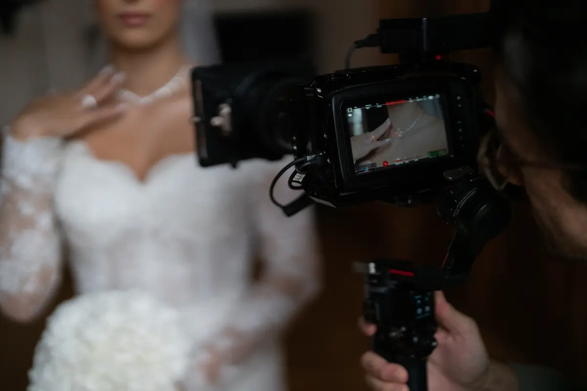 A camera operator films a bride in a white lace wedding dress holding a bouquet, capturing details of her jewelry.