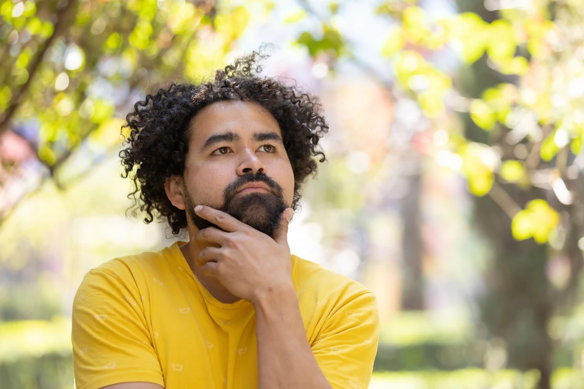 A man with curly hair wearing a yellow shirt sits in a park, resting his chin on his hand while deep in thought, surrounded by lush greenery.