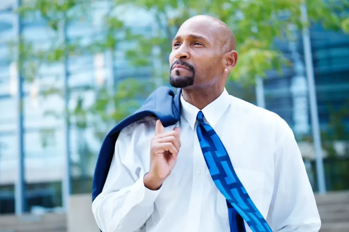 A mature businessman in a white shirt and blue tie gazes thoughtfully into the distance, with a blue jacket slung over his shoulder, standing outside a modern building with glass windows.