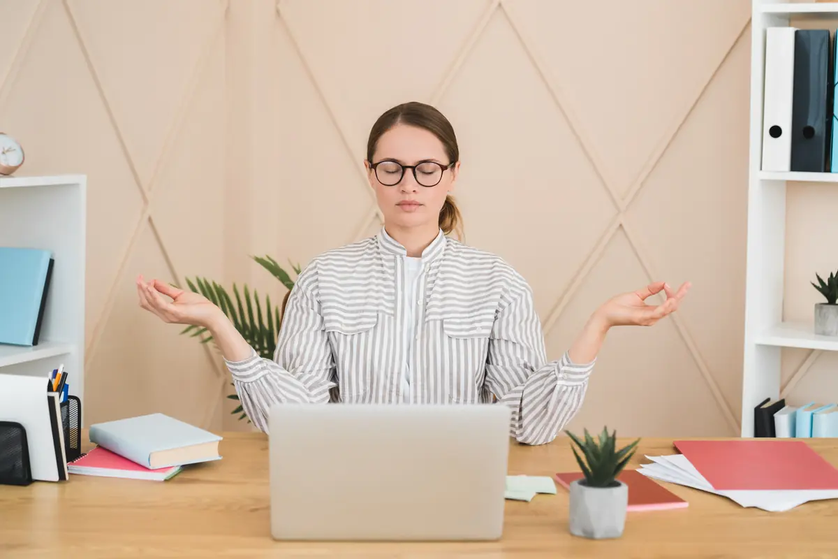 A woman sits at a desk with her eyes closed, meditating calmly before making a mindful financial decision, surrounded by books and plants.