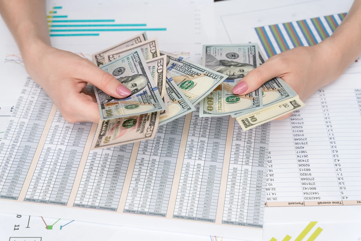 Hands holding various denominations of U.S. dollar bills over financial charts and graphs spread on a table.