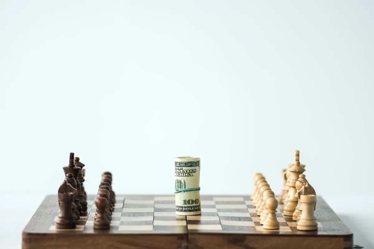Chess pieces arranged on a board with a rolled hundred-dollar bill standing upright in the center, symbolizing strategy in financial decisions.