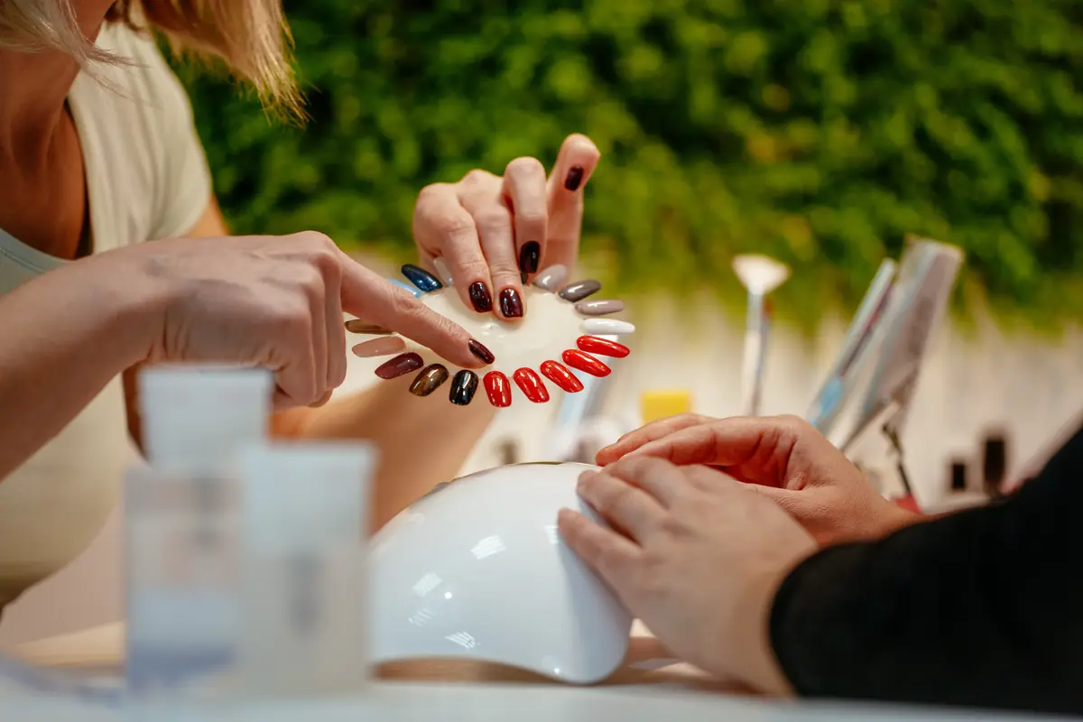 A person selecting a nail polish color from a palette in a nail salon, with another person's hand positioned under a UV nail lamp in a setting with a green plant background.