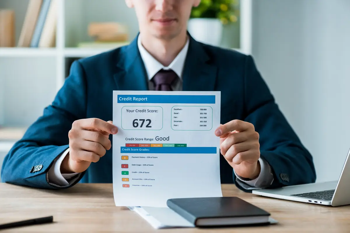 A person in a suit holding a credit report with a credit score of 672 displayed, indicating a "Good" credit score range, on a desk with a notebook and pen.