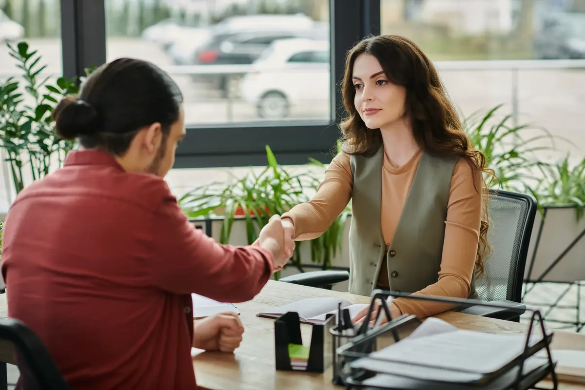 Two people sitting at a table in an office setting, engaged in a handshake, surrounded by paperwork and plants.