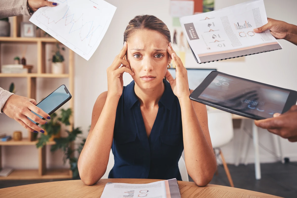 A woman sitting at a desk looks overwhelmed as she is surrounded by numerous hands holding charts, a smartphone, and a tablet.