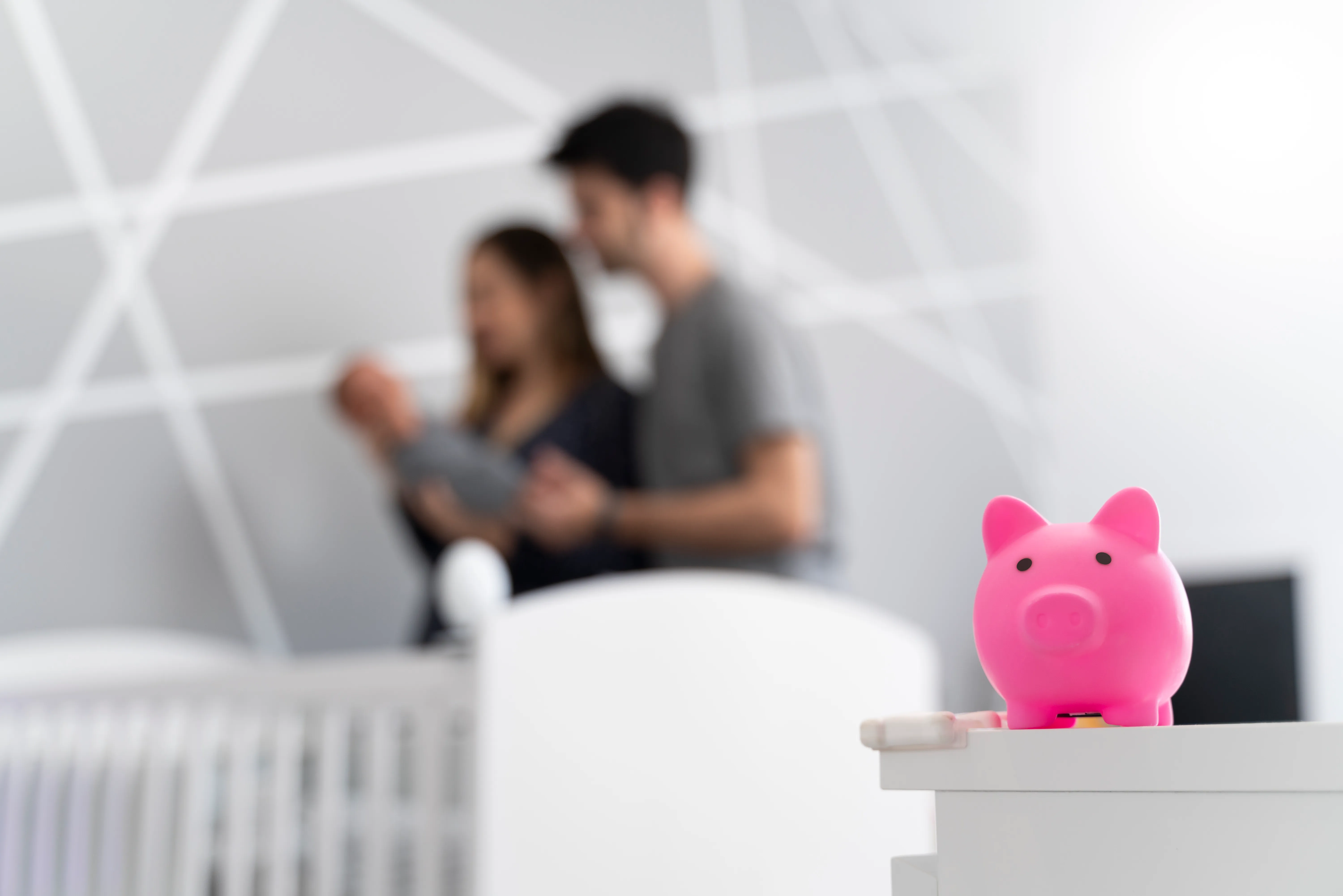 A pink piggy bank on a table in the foreground with a blurred background of a couple holding a baby near a crib.