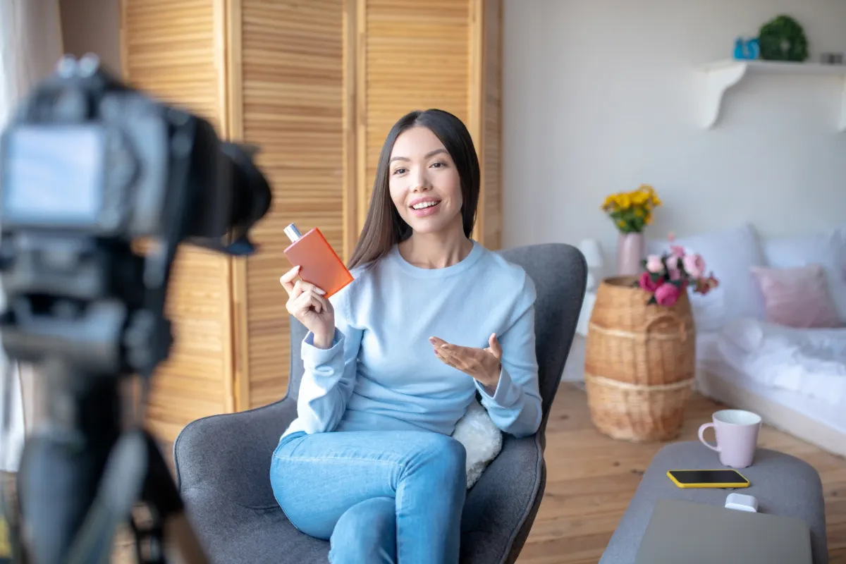 A woman seated in a cozy room holding a perfume bottle, speaking to a camera, with a warm, inviting background.