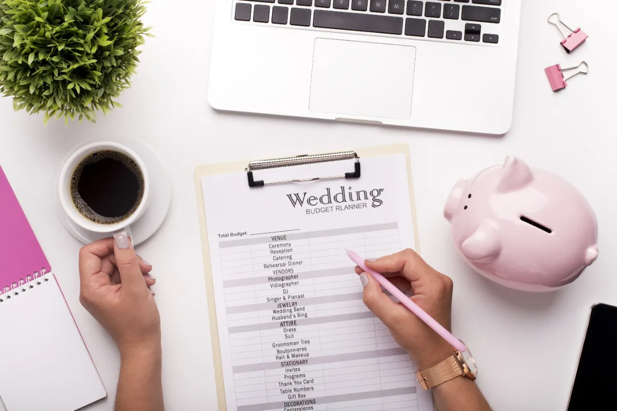 Hands writing on a wedding budget planner on a clipboard, surrounded by a laptop, a coffee cup, a pink piggy bank, and stationery supplies.