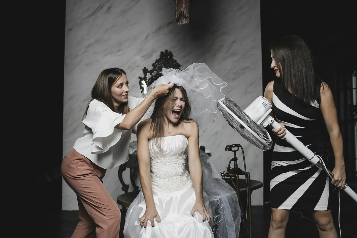 Bride laughing with her friends as one playfully adjusts her veil and another holds a standing fan, creating a fun and lively atmosphere during wedding preparations.