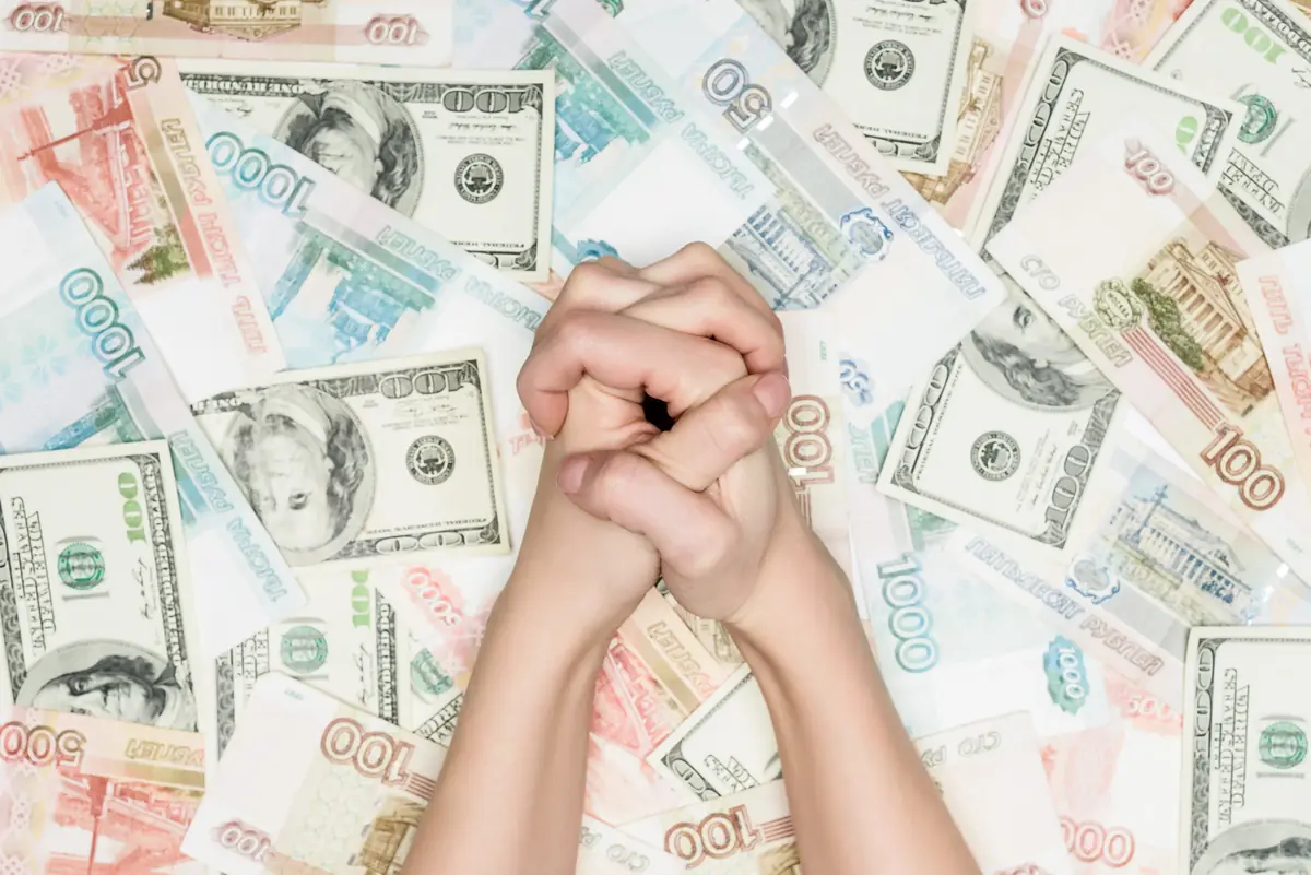 Hands clasped in a praying gesture over a scattered background of various international currency notes.
