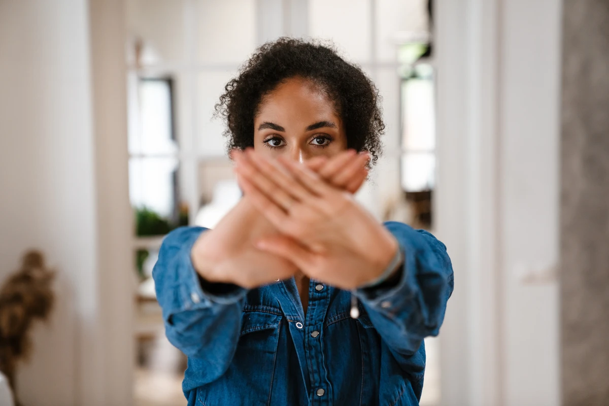 A person standing indoors crossing their arms in front of their face while looking directly at the camera, wearing a denim shirt.