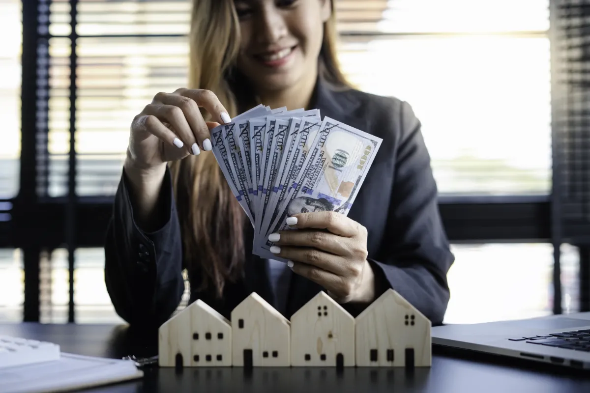 A person holding a stack of U.S. dollar bills above small wooden house models, symbolizing real estate investment.