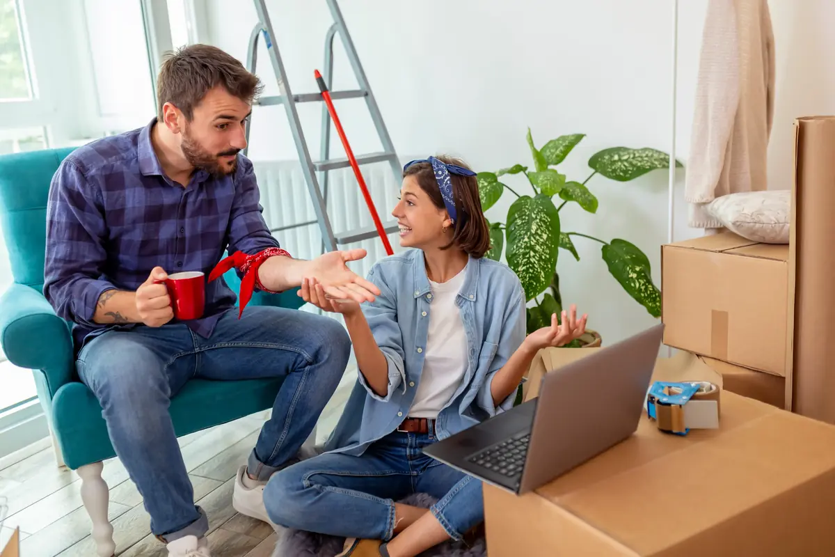 A couple sitting on the floor surrounded by moving boxes, discussing something while a laptop rests nearby, with a ladder and plant in the background.