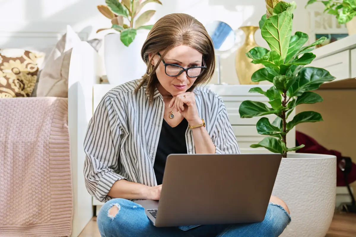 A woman wearing glasses and a striped shirt is seated on the floor, focused on her laptop, surrounded by plants in a bright indoor setting.