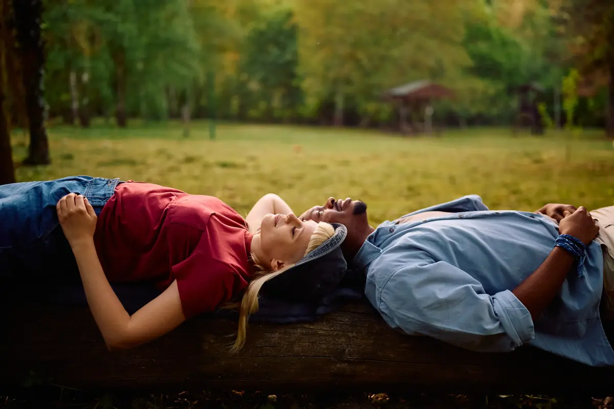 A couple lying back-to-back on a log, relaxing in a lush, green park under a canopy of trees.