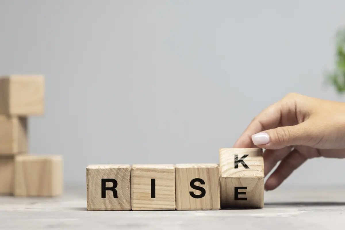 Wooden blocks spelling "RISE" are arranged on a surface, with a hand turning the last block to change the word to "RISK".