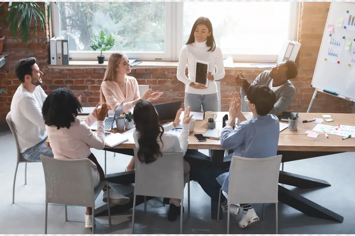 Seven colleagues sit around a conference table in a modern office, with a woman standing and holding documents as others applaud and engage in conversation.