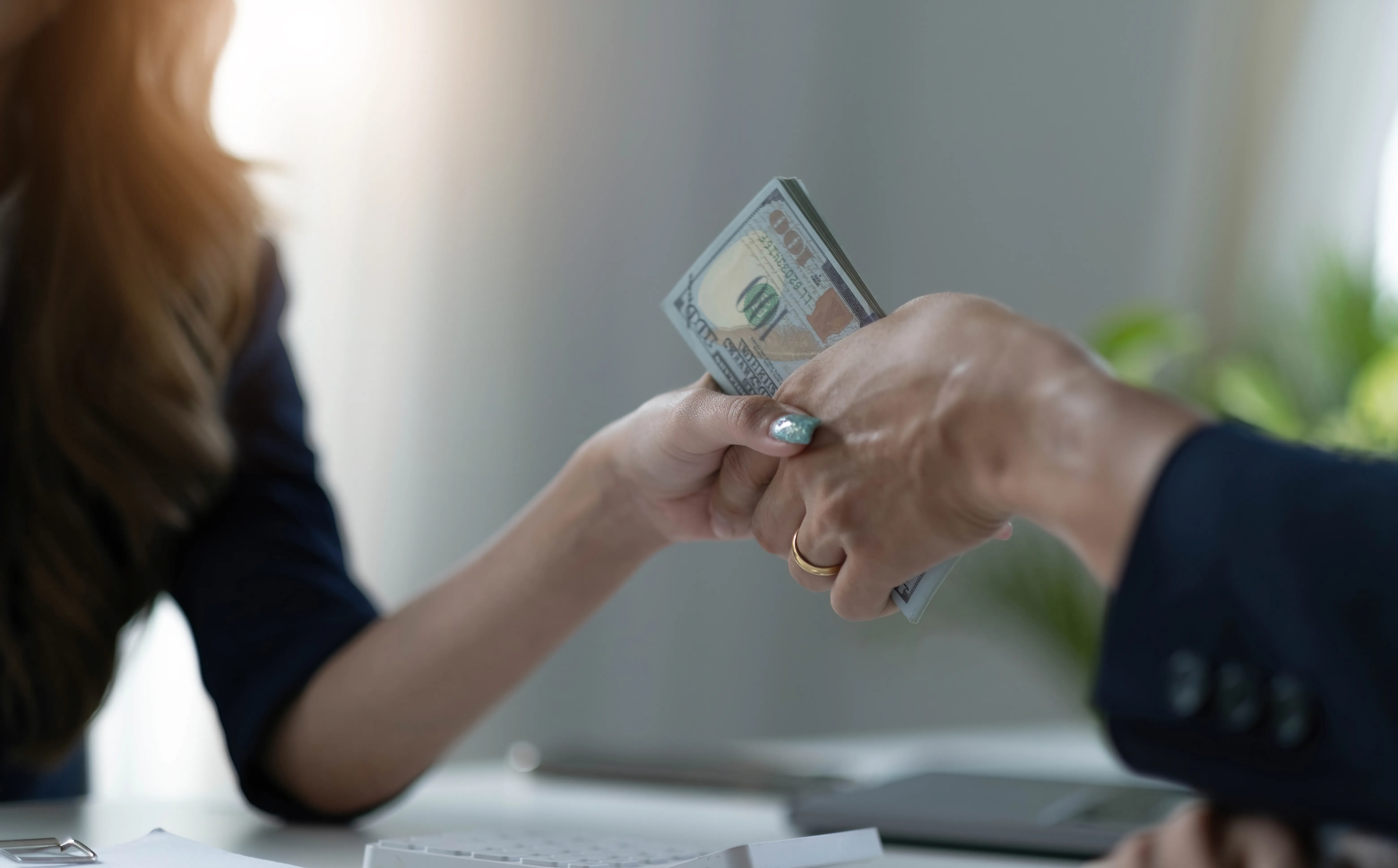 Two people exchanging a stack of hundred-dollar bills across a table, with visible hands and a blurred background.